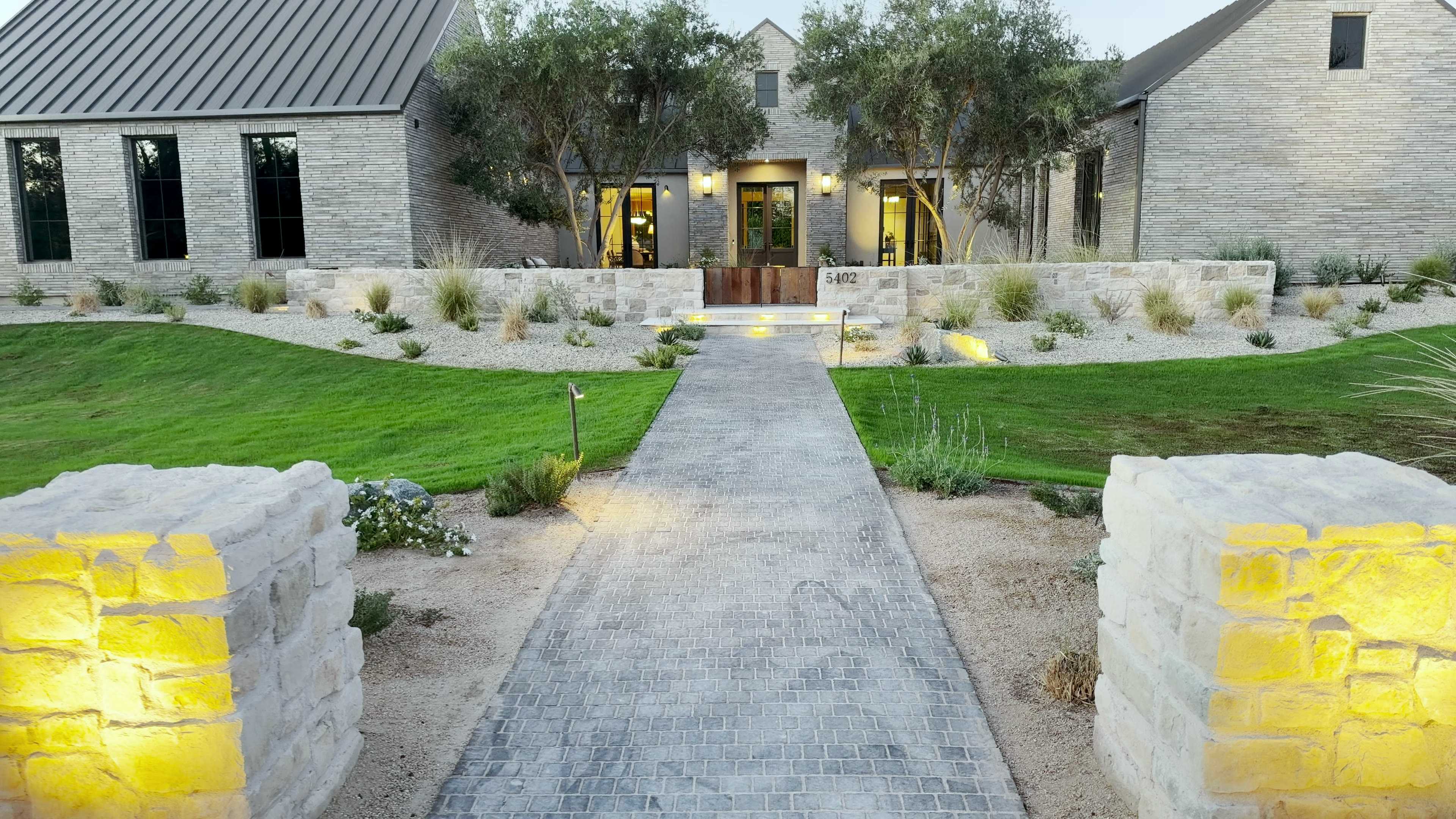 Front walk of a home in Arcadia at dusk, between lit stone pillars and across a lawn to the entry